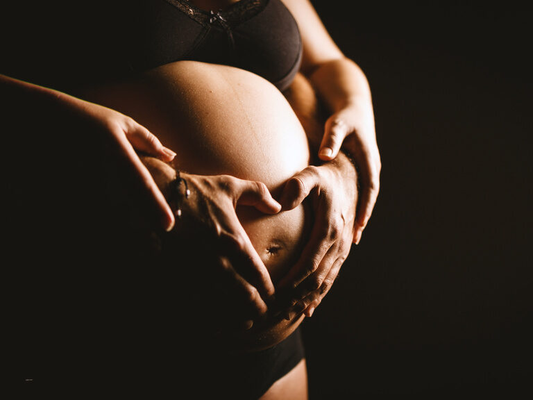 Hände umschlingen den Babybauch einer Frau von hinten.