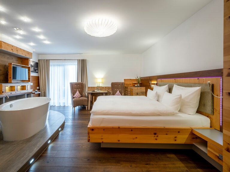 Modernes Zimmer mit Doppelbett mit LED-Beleuchtung, davor offene Badewanne, Flachbild-TV und Sitzecke