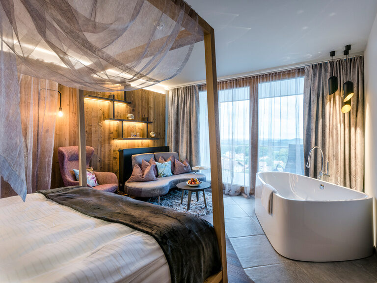 Modernes Zimmer mit Himmelbett, Badewanne und Balkon im Wellnesshotel Hüttenhof Modernes Zimmer mit Himmelbett, Badewanne, Sofa, Holzwände mit Beleuchtung und Balkon