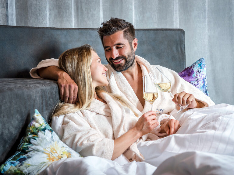Ein Paar in Bademänteln verbringt gemeinsam Zeit in einem schönen Rückzugsort und stößt mit einem Glas sekt an.